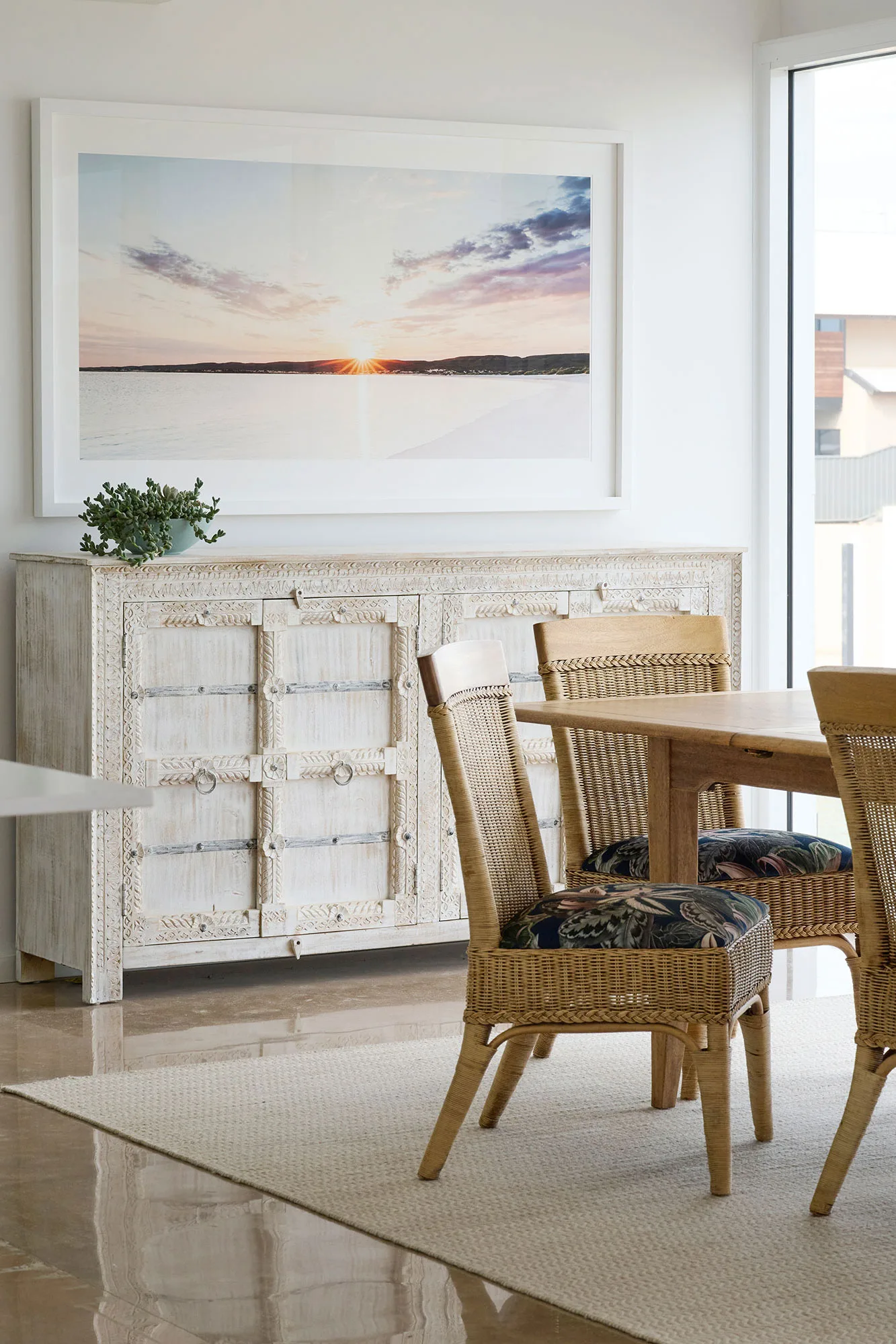 Exmouth I home — dining with carved white-washed sideboard, framed sunset photograph and rattan chairs